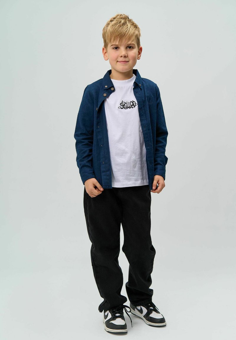 Cămașă albastră cu butoni peste un tricou alb cu grafică, pantaloni negri și adidași în alb și negru. Copilul stă în fața unui fundal deschis.