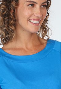 Blaues Oberteil mit weitem Ausschnitt und strukturierter Oberfläche. Das Model hat lockige Haare und ein sanftes Lächeln, das das Design des Ausschnitts zur Geltung bringt.