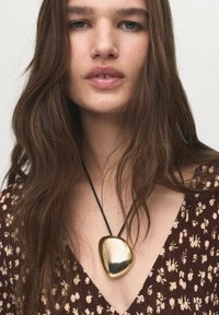 Pendentif en or avec une forme lisse et irrégulière sur un cordon noir, porté sur un haut à motif floral marron. Cheveux longs et ondulés.
