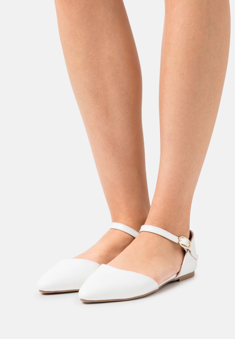 Białe płaskie buty z sztucznej skóry z ostrym noskiem i smukłym paskiem na kostkę z okrągłą złotą klamrą. Podeszwa jest płaska i brązowa.