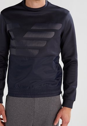 Homme portant un sweat-shirt bleu marine avec un grand logo ton sur ton d'aigle et un pantalon gris, debout devant un fond neutre.