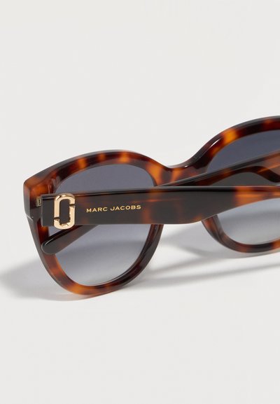 Καφέ γυαλιά ηλίου με σχέδιο χελώνας, με σκούρους φακούς και χρυσό λογότυπο Marc Jacobs στο πλαινό μέρος, σε ανοιχτό φόντο.