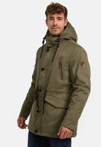 Parka verde oliva con capucha, presenta cordones negros, múltiples bolsillos y acentos marrones. Fabricada con una tela texturizada y duradera.