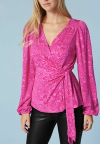 Blusa envoltura fucsia con mangas largas, textura floral, escote en V profundo y detalle de lazo en la parte delantera, combinada con pantalones de imitación de cuero negros.