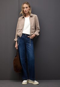 Helle beige Wildlederjacke mit Kordelzügen, weißem T-Shirt, blauen weit geschnittenen Jeans und hellen Sneakern. Eine braune Tasche ergänzt das Outfit.