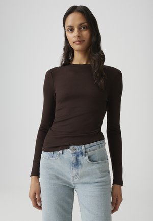 Vrouw met lang donker haar, gekleed in een aansluitend donkerbruin topje met lange mouwen en lichtblauwe hoog getailleerde spijkerbroek, staand tegen een effen achtergrond.