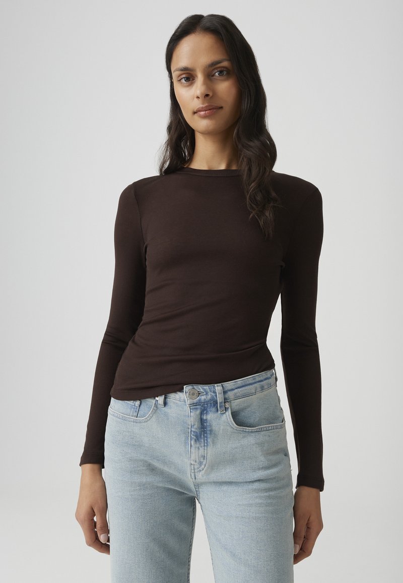 Vrouw met lang donker haar, gekleed in een aansluitend donkerbruin topje met lange mouwen en lichtblauwe hoog getailleerde spijkerbroek, staand tegen een effen achtergrond.