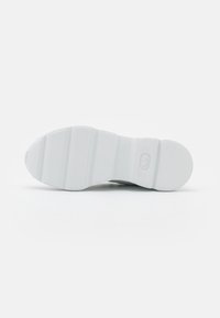Semelle de chaussure en caoutchouc blanc avec de profondes rainures horizontales et un petit logo ovale embossé près du talon sur un fond uni.