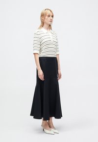 Woman wearing a white striped knit top, long black skirt, and white pointed low-heeled shoes standing against a plain background.