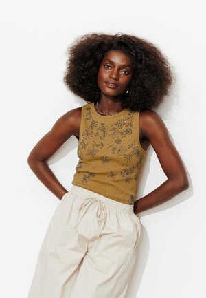 Femme aux cheveux bouclés et volumineux portant un haut olive sans manches avec broderie de perles et un pantalon ample clair à cordon, mains sur les hanches.