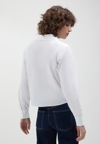 Blusa bianca a maniche lunghe con colletto, texture liscia, design cropped e polsini abbottonati, abbinata a jeans in denim scuri.