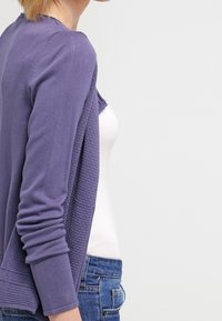 Cardigan tricoté violet avec un dos texturé, devant ouvert et manches longues, porté sur un haut blanc et un jean bleu.