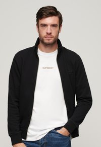 Sort lynlåsjakke med høj krave, båret over en hvid t-shirt med "SUPERDRY" i diskret broderi. Denimjeans er synlige.