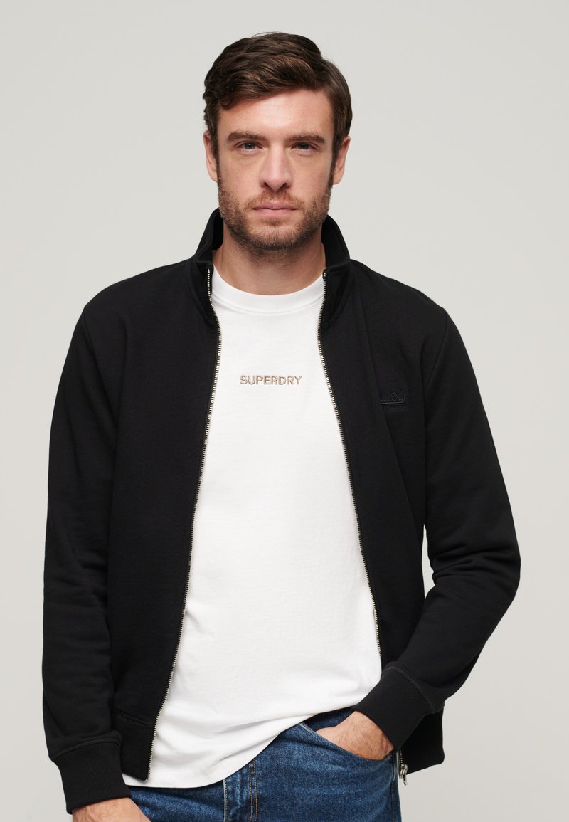 Sort lynlåsjakke med høj krave, båret over en hvid t-shirt med "SUPERDRY" i diskret broderi. Denimjeans er synlige.