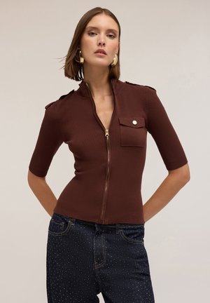 Bruine ribbelige zip-up top met korte mouwen, voorzien van een klepzak en gouden hardware-accenten. Gecombineerd met donkere jeans met kleine studs.
