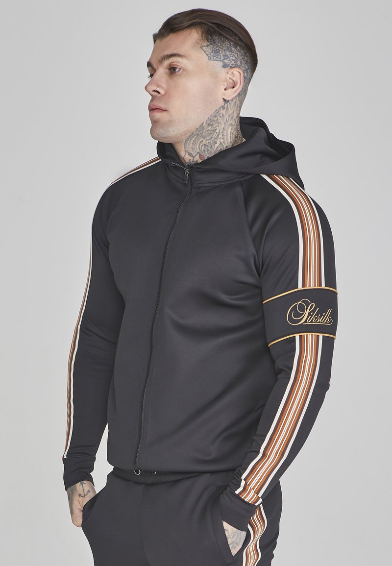 Sudadera con cremallera negra y mangas a rayas contrastantes en marrón, blanco y dorado. Presenta un logotipo bordado en la manga y un bolsillo frontal.