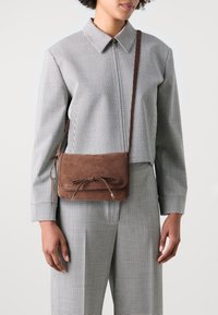 Brązowa zamszowa torebka na ramię o płaskim kształcie, z zapięciem na sznurek i srebrnymi akcentami; noszona z garniturem w kratę.