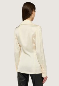 Camisa larga de satén color crema con mangas largas, cuello con muesca, costuras en los hombros, puños con botones y un ajuste entallado, combinada con pantalones de cuero negros.