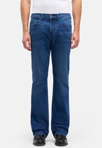 Dzwony z ciemnoniebieskiego denimu z pięcioma kieszeniami, metalowym zapięciem na guzik i kontrastującymi przeszyciami. Noszone z czarnymi butami.