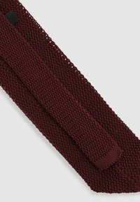 Cravat tricotat burgund cu un design texturat, cu țesătură deschisă. Prezintă o bandă solidă și netedă în centru și un capăt ascuțit.