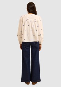 Blusa bianca ricamata con motivi floreali blu, maniche a sbuffo e un fit rilassato, abbinata a jeans a gamba larga di un blu scuro.
