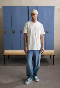 Ljusbeige bomullströja med en liten orange grafik, ihop med lösa blå denimjeans och teal sneakers, mot en bakgrund av blå skåp.