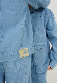 Jachetă din denim bleu deschis, cu logo brodat și patch. Detalii notabile includ un buzunar frontal și cusături vizibile.