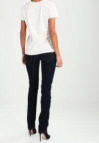 Kvinna iklädd en vit kortärmad skjorta, mörka tajta jeans och svarta högklackade skor, står vänd bort mot en vit bakgrund.