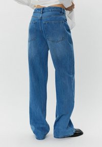 Široke plave traperice s visokim strukom, dva stražnja džepa i suptilnom izbljeđenom teksturom. Rubovi su malo rastrgani.