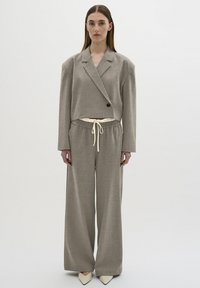 Beiger Cropped-Blazer mit einem einzelnen schwarzen Knopf, kombiniert mit weiten beigen Hosen mit elastischem Bund und weißem Kordelzug. Spitz zulaufende Absatzschuhe.