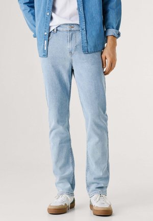 Mann trägt hellblaue Jeans, weiße Sneaker mit braunen Sohlen, weißes T-Shirt und offenes blaues Jeanshemd und steht vor einem einfarbigen Hintergrund.