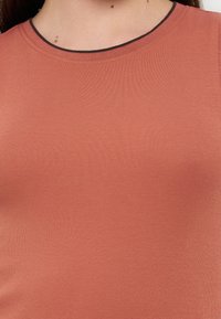 Κοντινό πλάνο ενός ατόμου που φοράει ριμπ, χωρίς μανίκια τοπ σε τερακότα χρώμα με λεπτό μαύρο περίγραμμα γύρω από το λαιμόκοψη scoop.