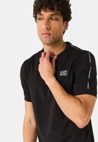 Camiseta negra de manga corta con escote redondo y logo en el pecho. Presenta el branding en blanco a lo largo de los hombros y una textura de tela suave.