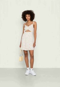 Vestido de verão floral em cores claras com um design recortado, alças de ombro ajustáveis e uma saia rodada. Combinado com ténis brancos e uma pequena mala amarela.