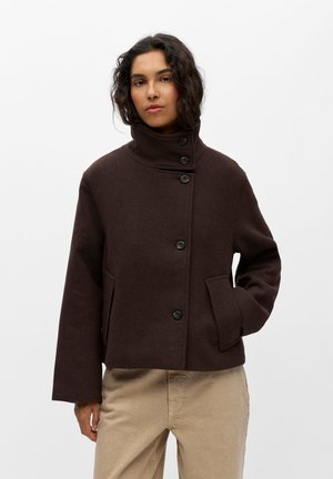 Kvinde iført en mørkebrun uldjakke med knapper og høj krave og beige bukser, stående med den ene hånd i lommerne mod en hvid baggrund.