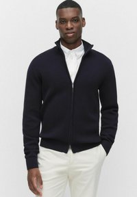 Marinblå zip-up tröja med hög krage, ribbade muddar och nederkant. Bärs över en vit skjorta med krage, tillsammans med ljusfärgade byxor.