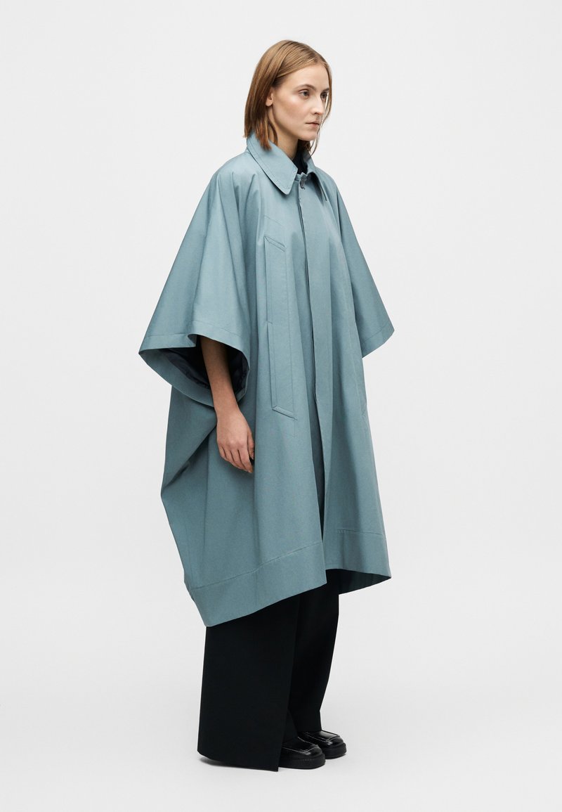 Manteau style poncho bleu clair, oversize, avec col, poches latérales et coupe décontractée. Porté sur un pantalon noir à jambes larges et des chaussures.