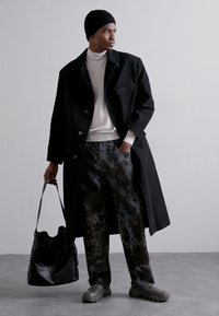 Black long coat, cream turtleneck, dark patterned trousers, gray shoes. Accessories include a textured black bag and a knitted black beanie.