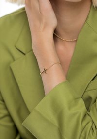 Zlata zapestnica z verižico in obeskom križa, nošena na goli zapestju. V ozadju zeleno tkaninsko suknjič prikazuje gladko teksturo in krojen a prilegajoč se kroj.