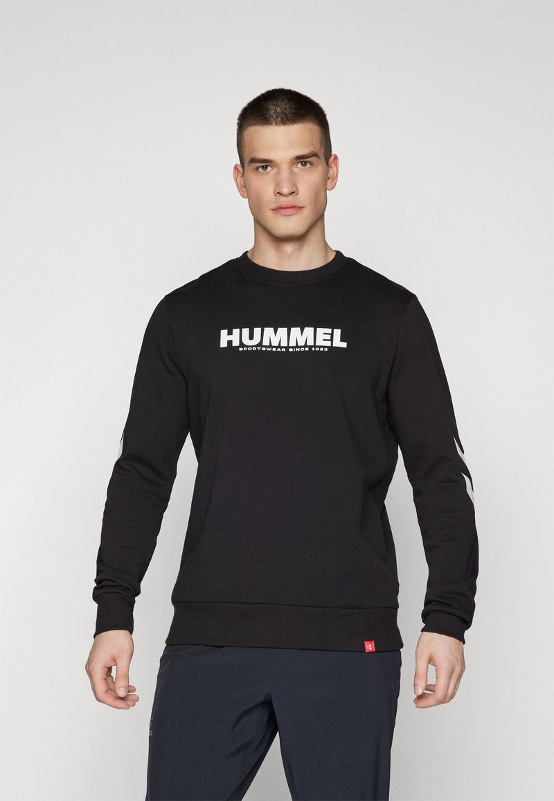 Svart hettegenser med lange ermer, med en dristig hvit "HUMMEL"-logo og liten rød etikett, laget av myk stoff med vanlig passform.
