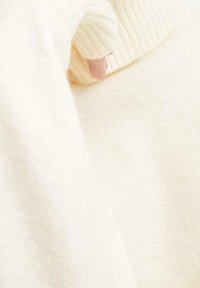 Ruka s částečně viditelným palcem uvnitř manžety krémově barevného žebrovaného pleteného svetru.