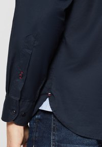 Poignet de chemise bleu marine à manches longues avec détail de bouton rouge, porté sur un jean bleu foncé, montrant une couture latérale avec une petite broderie tricolore.