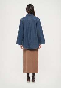 Jaqueta de ganga com um corte relaxado, mangas largas e detalhe de pregas nas costas. Usada sobre uma saia longa castanha, com uma textura suave e cor neutra.