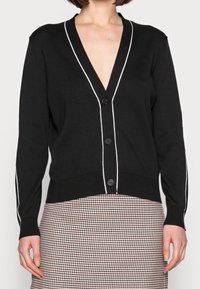 Svart kavaj med djup V-ringning, vit kantning längs kanterna, fem knappar och ribbade manschetter. Bärs över en mönstrad kjol.