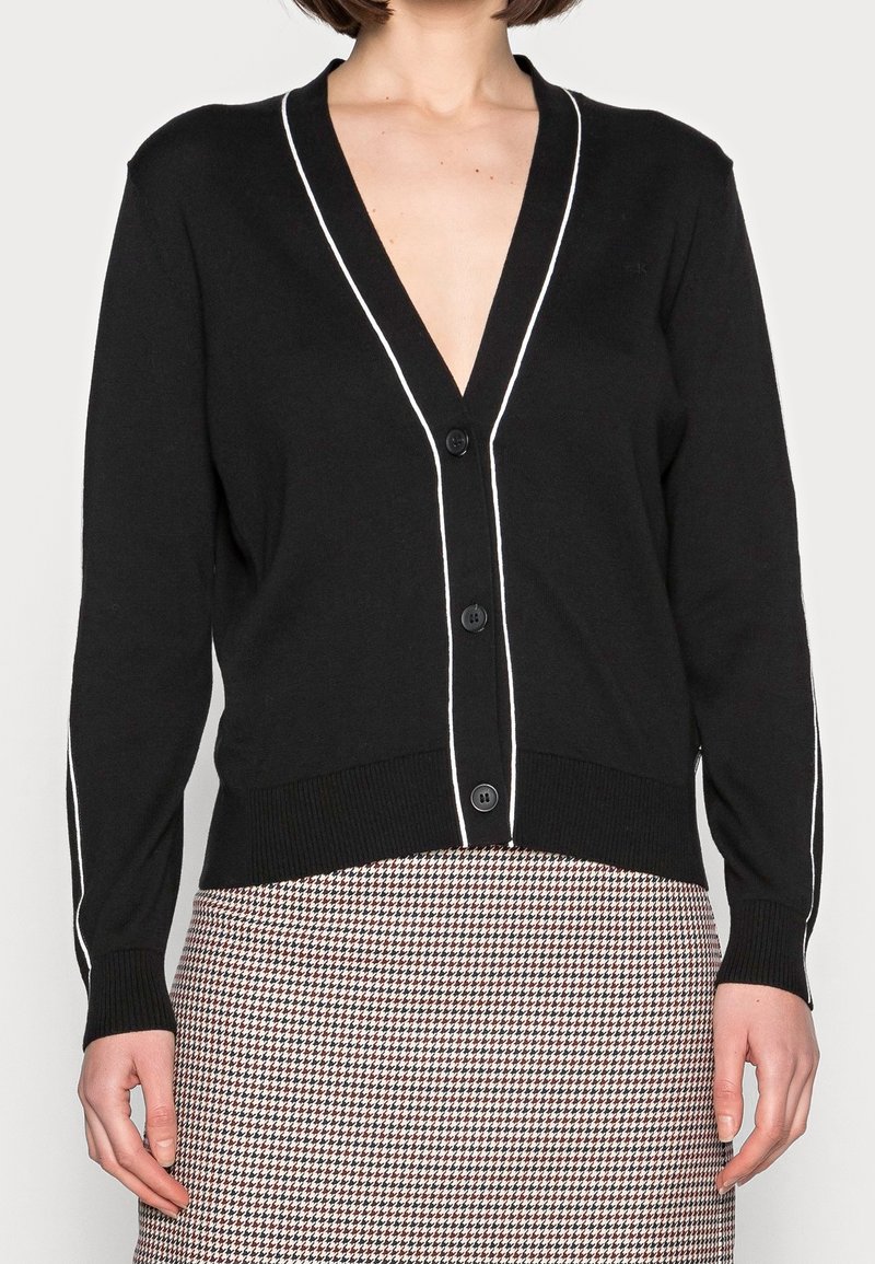 Svart kavaj med djup V-ringning, vit kantning längs kanterna, fem knappar och ribbade manschetter. Bärs över en mönstrad kjol.
