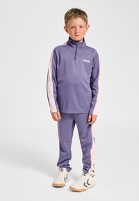 Lila Sport-Trainingsanzug mit langen Ärmeln, Halb-Reißverschluss am Kragen und rosa seitlichen Akzenten. Verfügt über eine glatte Textur und ein Logo auf der Brust. Weiße Turnschuhe.