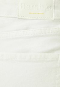 Vitt denimtyg med en slät yta, försett med en etikett som har texten "Bershka Denim Collection" i präglade bokstäver med guldaccenter.