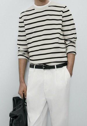 Homme portant une chemise blanche à manches longues avec des rayures horizontales noires, pantalon blanc, ceinture noire, tenant un sac en cuir noir.