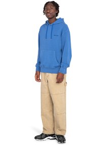 Sudadera de algodón azul con bolsillo frontal, capucha con cordones y logo. Combinada con pantalones de pierna ancha beige. Zapatillas negras con diseño texturizado.