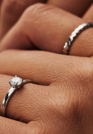 Zilveren ring met een ronde diamanten siersteen, gezet in een vierpoots zetting, naast een glazen zilveren band op een hand.